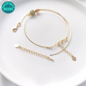 Op de afbeelding: Gouden armband met een groene stenen kraal en een hartvormige hanger. De armband is gemaakt van twee dunne draden en heeft een karabijn sluiting. De hartvormige hanger is bevestigd aan een ketting.