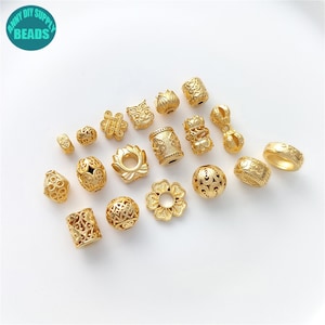 Könnte beinhalten: Eine Sammlung von 18 goldfarbenen Metallperlen mit filigranen Designs. Die Perlen sind in verschiedenen Formen und Größen, darunter rund, oval und zylindrisch. Einige Perlen haben florale Muster, während andere geometrische Designs aufweisen.