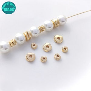 May include: Gold-toned metal beads in various shapes, including round, oval, and triangular, with a hole in the center. The beads are scattered on a white surface.