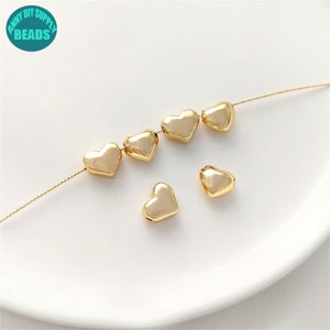 May include: A string of gold heart-shaped beads, with a few extra beads scattered on a white surface. The beads have a slightly iridescent finish.