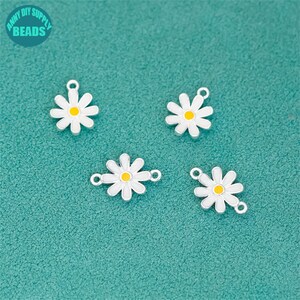K&ouml;nnte beinhalten: Vier charmartige Anh&auml;nger in Form von G&auml;nsebl&uuml;mchen mit wei&szlig;en Bl&uuml;tenbl&auml;ttern und gelben Zentren. Die Anh&auml;nger haben einen silberfarbenen Rand und eine Schlaufe zum Befestigen an Schmuck. Der Hintergrund ist eine t&uuml;rkisfarbene Oberfl&auml;che. Der Text "ANNY DIY SUPPLY BEADS" befindet sich in der oberen linken Ecke.