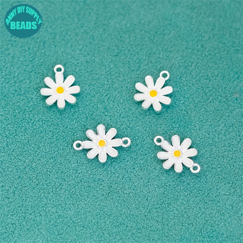K&ouml;nnte beinhalten: Vier charmartige Anh&auml;nger in Form von G&auml;nsebl&uuml;mchen mit wei&szlig;en Bl&uuml;tenbl&auml;ttern und gelben Zentren. Die Anh&auml;nger haben einen silberfarbenen Rand und eine Schlaufe zum Befestigen an Schmuck. Der Hintergrund ist eine t&uuml;rkisfarbene Oberfl&auml;che. Der Text "ANNY DIY SUPPLY BEADS" befindet sich in der oberen linken Ecke.