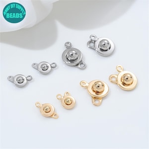 Könnte beinhalten: Eine Auswahl an Metall-Schmuckverschlüssen in Silber- und Goldtönen. Die Verschlüsse haben ein rundes, knopfartiges Design mit einer kleinen Schlaufe auf jeder Seite zur Befestigung. Das Bild enthält den Text "ANNY DIY SUPPLY BEADS" in einem kreisförmigen Logo.