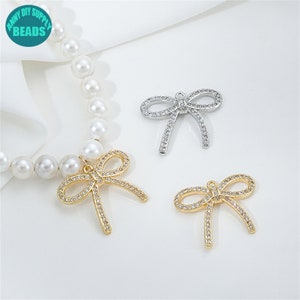 Pode incluir: Três pingentes de laço em prata e ouro com strass transparentes. Os pingentes estão sobre um fundo branco.