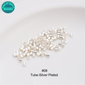 May include: A close-up of a pile of silver-plated tube beads. The beads are small and round, with a smooth finish. The beads are arranged in a random pattern, and some of the beads are overlapping. The beads are on a white background. #08 Tube-Silver Plated