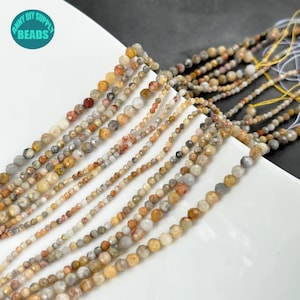 Puede incluir: Una hebra de cuentas de piedras preciosas facetadas en tonos marrones, beige y blancos. Las cuentas son pequeñas y redondas, y están enhebradas en un hilo transparente. Las cuentas están dispuestas en un patrón suelto y fluido.