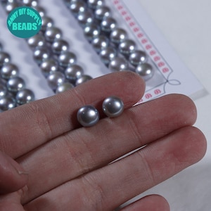 Può includere: Due perle rotonde color grigio argento, di circa 10 mm di diametro, sono tenute in una mano. Le perle sono lisce e hanno un leggero bagliore iridescente.