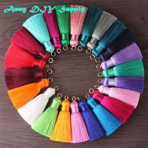 Puede incluir: Surtido de 24 borlas de seda de colores con anillas de salto doradas. Las borlas están dispuestas en círculo sobre una superficie de madera.