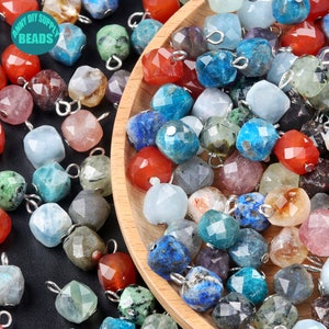 May include: Assortment of faceted gemstone beads with silver jump rings. The beads are in various colors, including blue, green, red, and purple. The beads are in a wooden bowl.