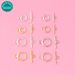 May include: Twelve silver and gold toggle clasps with a round ring and a straight bar. The clasps are arranged in four rows of three on a pink background.