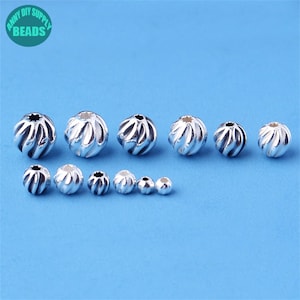 Puede incluir: Un conjunto de cuentas plateadas de varios tamaños, con un diseño en espiral, sobre un fondo azul. Las cuentas son probablemente para la creación de joyas. La imagen incluye el texto "ANNY DIY SUPPLY BEADS".