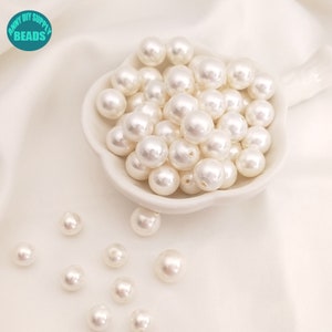 May include: A white bowl filled with white pearl beads. The bowl is on a white surface. There are several white pearl beads scattered around the bowl.