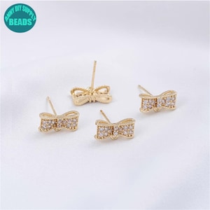 May include: Gold-colored bow-shaped stud earrings with clear crystal accents. The earrings are displayed on a white surface. The image also includes a teal circle with the text "ANNY DIY SUPPLY BEADS".