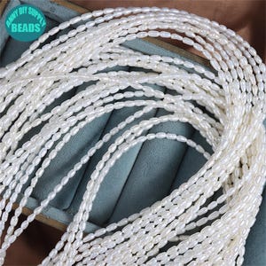 K&ouml;nnte beinhalten: Nahaufnahme von mehreren Str&auml;ngen wei&szlig;er, oval geformter Perlen. Die Perlen sind aufgef&auml;delt und erzeugen einen Kaskadeneffekt. Das Bild enth&auml;lt den Text "ANNY DIY SUPPLY BEADS" in einem kreisf&ouml;rmigen Logo. Wahrscheinlich S&uuml;&szlig;wasserperlen.
