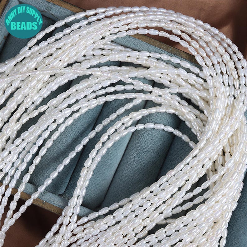 K&ouml;nnte beinhalten: Nahaufnahme von mehreren Str&auml;ngen wei&szlig;er, oval geformter Perlen. Die Perlen sind aufgef&auml;delt und erzeugen einen Kaskadeneffekt. Das Bild enth&auml;lt den Text "ANNY DIY SUPPLY BEADS" in einem kreisf&ouml;rmigen Logo. Wahrscheinlich S&uuml;&szlig;wasserperlen.