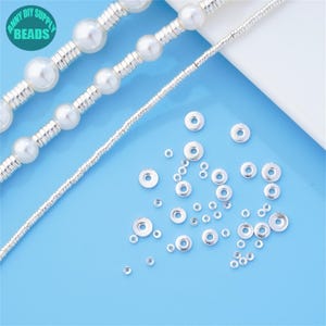 Puede incluir: Primer plano de tapas de cuentas de metal plateadas esparcidas sobre una superficie azul claro. Las tapas de cuentas son redondas con una parte superior plana y un borde elevado. También hay tres hebras de cuentas plateadas y perlas blancas en el fondo.