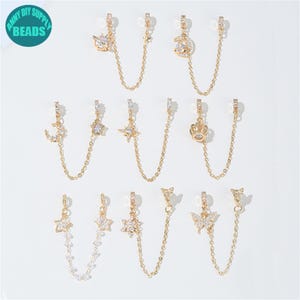 May include: Gold-toned double-piercing earrings with various charms, including stars, moons, crowns, and butterflies. Each earring features a delicate chain connecting two decorative elements, all adorned with sparkling crystals. The earrings are displayed on a white background.