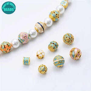 Puede incluir: Una colección de cuentas de esmalte coloridas en varias formas y tamaños, incluyendo diseños redondos y florales. Las cuentas presentan detalles dorados y están ensartadas con perlas blancas. La imagen incluye el texto "ANNY DIY SUPPLY BEADS".