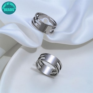 Op de afbeelding: Twee zilverkleurige metalen ringen met een breed bandontwerp, geometrische uitsparingen aan de zijkanten en een glad, massief middengedeelte. De ringen hebben een gepolijste, reflecterende afwerking en een open structuur.