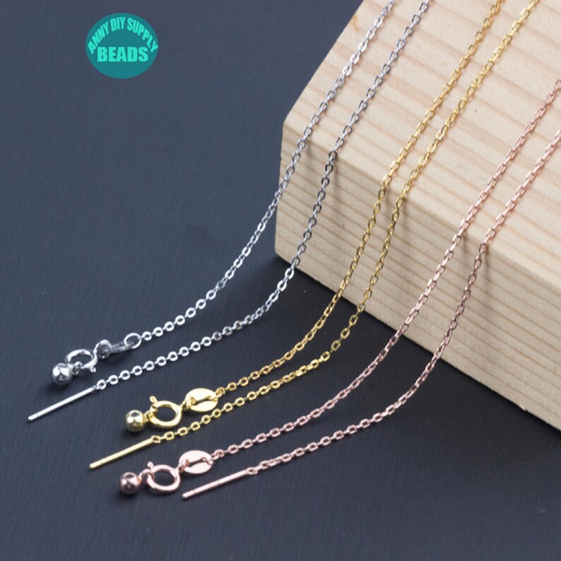 May include: Four silver, gold, and rose gold chains with a lobster clasp and a small ball on the end. The chains are laying on a wooden surface.