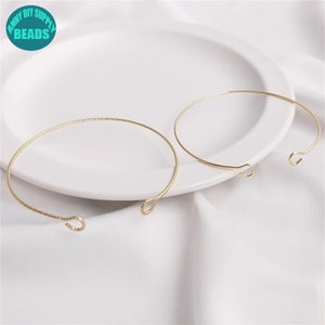Peut inclure: Deux bracelets en métal doré de conception simple et ouverte. L'un des bracelets présente un motif texturé, tandis que l'autre est lisse. Les bracelets sont présentés sur une assiette blanche, avec le texte "ANNY DIY SUPPLY BEADS" dans un cercle turquoise en haut à gauche.