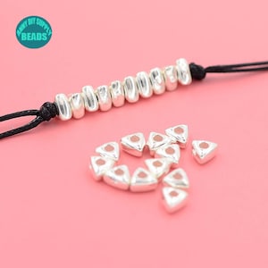 Pode incluir: Contas de metal prateado em uma variedade de formas, incluindo redondas e triangulares. As contas estão dispostas sobre uma superfície rosa. As contas redondas estão enfiadas em um cordão preto. As contas triangulares estão espalhadas ao redor das contas redondas.