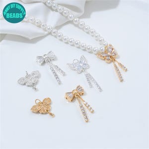 Op de afbeelding: Een verzameling sieradenbedels in vlinder- en strikvormen. De bedels zijn goud- en zilverkleurig, met heldere kristallen accenten. Op de achtergrond is een parelketting te zien. De afbeelding bevat een turquoise cirkel met de tekst "ANNY DIY SUPPLY BEADS".