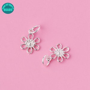 Könnte beinhalten: Zwei silberne Blumenanhänger mit einer Öse oben zum Anbringen an Schmuck. Die Blumen haben eine runde Mitte mit einer strukturierten Oberfläche und sechs Blütenblättern.