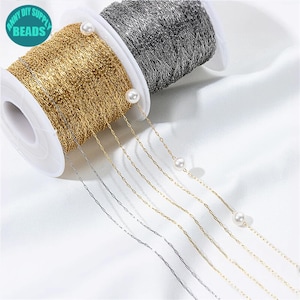 Puede incluir: Carretes de cadena dorada y plateada con perlas de imitación adjuntas. La cadena dorada está en un carrete blanco con el texto "ANNY DIY SUPPLY BEADS" en un círculo turquesa. Las cadenas se muestran sobre un fondo de tela blanca.