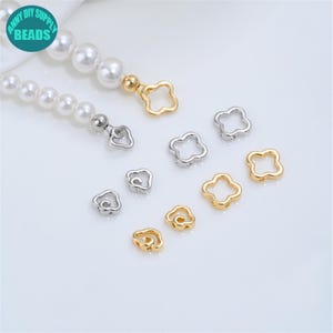 May include: A collection of gold and silver-toned metal clover-shaped beads and clasps, some attached to a strand of white faux pearls. The image includes a circular teal logo with the text ANNY DIY SUPPLY BEADS in the upper left corner.