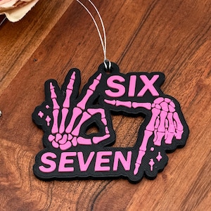 May include: A black air freshener with the words "SIX SEVEN" in pink, along with a hand gesture made of pink skeleton bones. The air freshener hangs from a silver string. A pink and cream-colored flower is in the background.