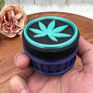 May include: A black and blue grinder with a teal marijuana leaf design on the top. The grinder has a textured bottom and a smooth top.