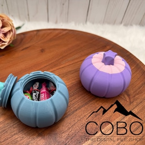 May include: A blue 3D-printed container with a lid, filled with sweets, sits open on a wooden tray. A second, closed container in purple and pink is next to it. The containers are pumpkin-shaped.
