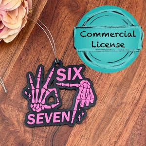 May include: Black air freshener with a pink skeleton hand design spelling out "SIX SEVEN". The air freshener has a commercial license tag and is hanging by a silver string. The background is a dark wood surface.