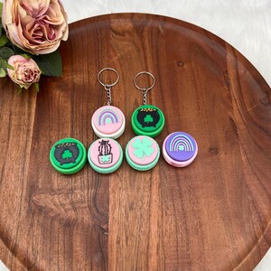 May include: Six round keychains with different designs. The keychains are green, pink, and purple with images of a pot of gold, a four-leaf clover, a rainbow, a cactus, and a pot of gold. The keychains are on a wooden surface.