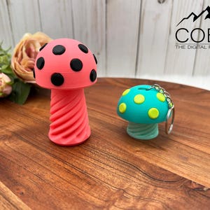 May include: A pink mushroom-shaped object with black spots and a spiral stem, alongside a smaller teal mushroom-shaped keychain with yellow spots and a silver metal ring. The text COBO THE DIGITAL FILE SHOP is visible in the upper right corner.