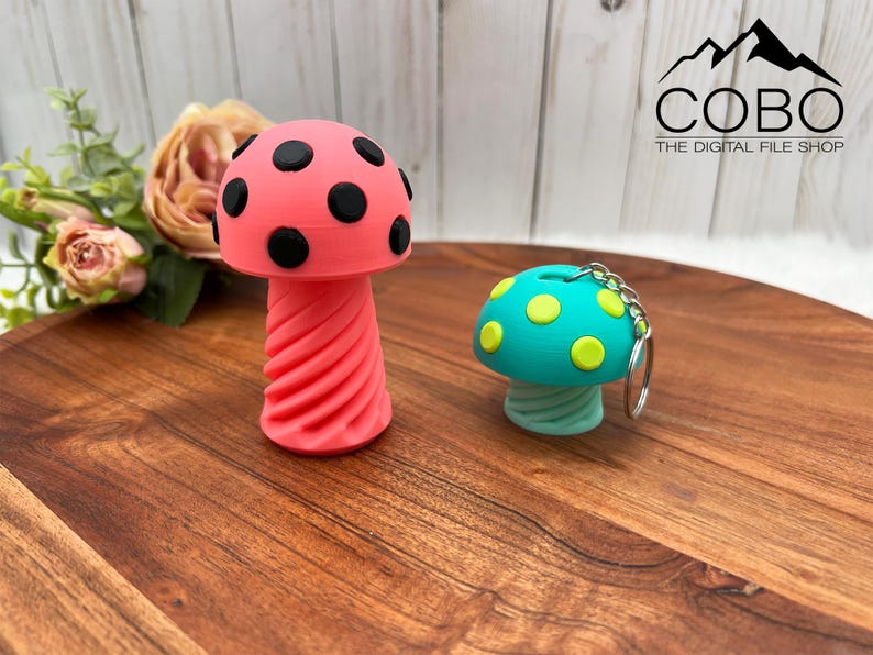 May include: A pink mushroom-shaped object with black spots and a spiral stem, alongside a smaller teal mushroom-shaped keychain with yellow spots and a silver metal ring. The text COBO THE DIGITAL FILE SHOP is visible in the upper right corner.