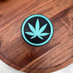 May include: A round wooden coaster with a teal blue and black marijuana leaf design.