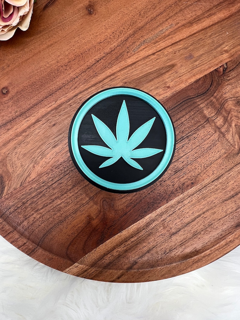 May include: A round wooden coaster with a teal blue and black marijuana leaf design.