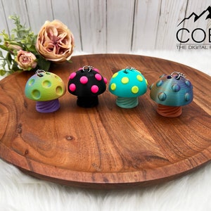 May include: Four mushroom-shaped keychains with metal rings. The first is green and yellow with a purple stem, the second is black with pink spots, the third is teal with yellow spots, and the fourth is iridescent blue with an orange stem. COBO THE DIGITAL FILE SHOP.
