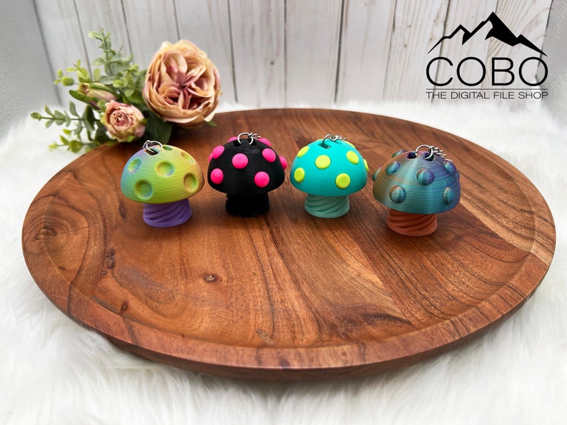 May include: Four mushroom-shaped keychains with metal rings. The first is green and yellow with a purple stem, the second is black with pink spots, the third is teal with yellow spots, and the fourth is iridescent blue with an orange stem. COBO THE DIGITAL FILE SHOP.