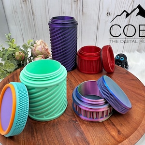 Op de afbeelding: Een verzameling kleurrijke, 3D-geprinte containers met een textuuroppervlak en schroefdeksels. De containers zijn in verschillende kleuren, waaronder paars, rood, groen en iriserend. De containers worden op een houten dienblad gepresenteerd.