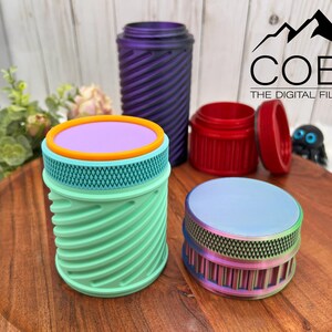 Op de afbeelding: Een verzameling kleurrijke, 3D-geprinte containers in verschillende maten en ontwerpen. De containers omvatten een turquoise en oranje cilindrische pot, een paarse spiraalpot, een rode pot met een deksel en een regenboogkleurige pot. Het COBO-logo is zichtbaar.