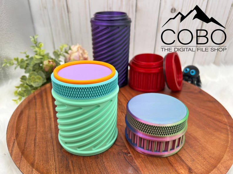 Op de afbeelding: Een verzameling kleurrijke, 3D-geprinte containers in verschillende maten en ontwerpen. De containers omvatten een turquoise en oranje cilindrische pot, een paarse spiraalpot, een rode pot met een deksel en een regenboogkleurige pot. Het COBO-logo is zichtbaar.