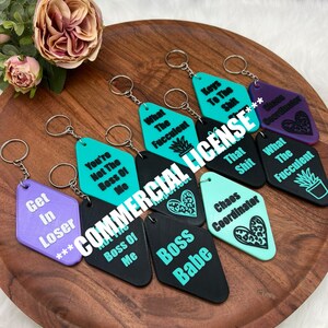 May include: A collection of novelty keychains in various colors, including black, teal, and purple. Each keychain features a unique phrase or design, such as "Boss Babe" and "Chaos Coordinator". The keychains are displayed on a wooden tray.