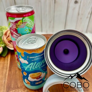 May include: Three aluminium cans with vibrant designs. One can is red and green with "Cherry Twist" text. Another is blue and orange with "Dream Float" and "Alani" text. A purple-lined tumbler is also present.