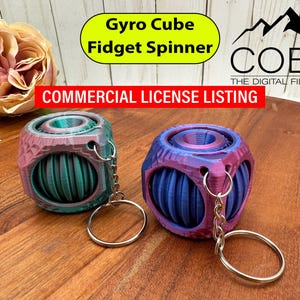 Puede incluir: Dos Gyro Cube Fidget Spinners multicolores con llaveros están sobre una superficie de madera. Los spinners tienen forma de cubo con un elemento giratorio central y un diseño colorido. La imagen incluye el texto "Gyro Cube Fidget Spinner" y "Commercial License Listing."