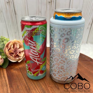 May include: A red, teal, and white Alani Nu Cherry Twist energy drink can next to a light blue can cooler with a silver leopard print design. The cooler holds a smaller can with an orange and blue design.