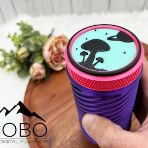 Puede incluir: Vaso reutilizable morado con tapa rosa y un diseño azul y negro de setas y un planeta en la tapa.