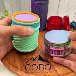 Könnte beinhalten: Eine Sammlung farbenfroher, zylindrischer Grinder mit strukturierten Seiten und Deckeln. Einer wird in der Hand gehalten und zeigt einen mintgrünen Körper mit einem lila und orangefarbenen Deckel. Ein anderer ist irisierend mit einem hellblauen Deckel. Das Logo "COBO" ist sichtbar.